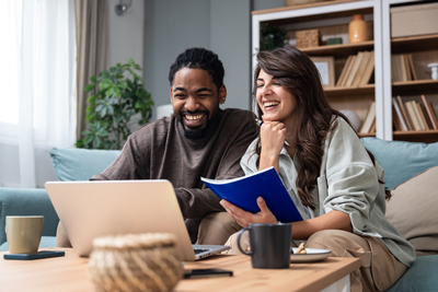 A man and woman sitting together on a couch, laughing and looking at a laptop, while the woman holds a notebook, in a cozy living room setting with plants and bookshelves.