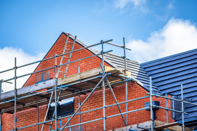 Scaffolding around a partially constructed brick house with a gray roof under a blue sky.