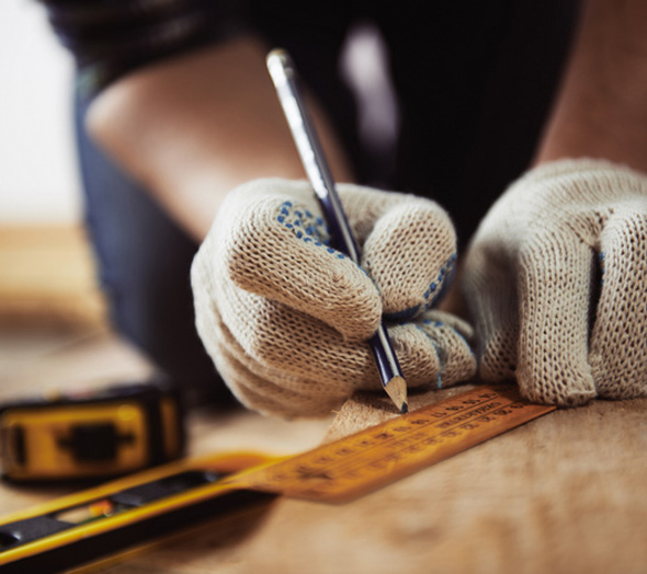 Person wearing gloves measuring and marking wood with a pencil, alongside a measuring tape, focusing on precision in a woodworking project.