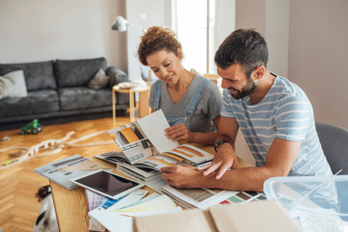 A couple sitting at a table covered with design materials, discussing home renovation ideas while looking through color swatches and samples in a bright, modern living room.