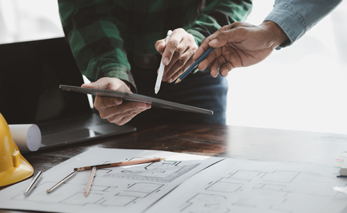 two hands holding a tablet with design plans and architectural sketches on a table surrounded by pens and a construction helmet