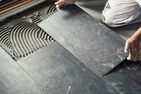 Person installing dark stone tiles on a textured adhesive surface, showcasing a flooring project.