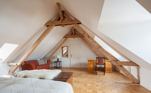 A cozy attic room featuring exposed wooden beams, a red sofa, a wooden desk, and a patterned chair, with natural light streaming in from the windows. Ideal for a relaxing retreat or workspace.