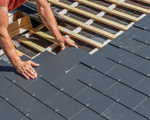 person installing dark gray slate roofing tiles on a wooden framework