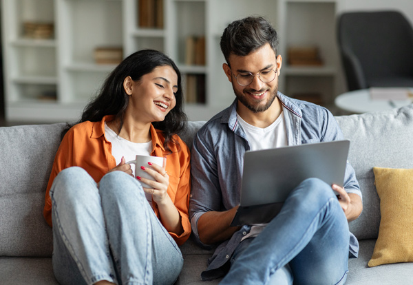 Couple sitting on a sofa, enjoying time together while looking at a laptop, with one holding a cup, conveying a relaxed and cozy atmosphere.