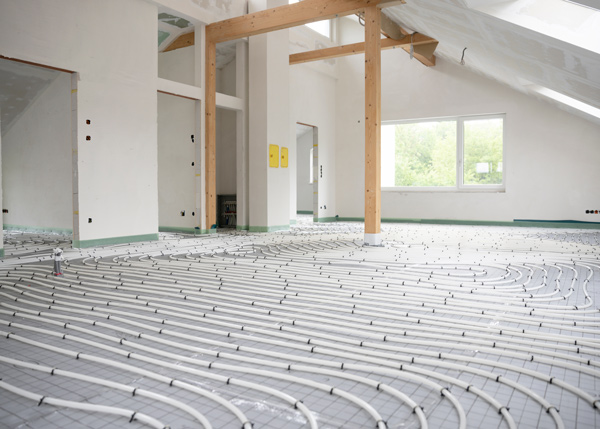 Interior view of a room under construction, featuring a floor installation with white tubing arranged in curved patterns, surrounded by exposed walls and wooden beams, indicating preparation for floor heating systems.