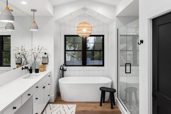 Modern bathroom with a freestanding tub, elegant lighting fixture, black fixtures, and a glass shower enclosure, featuring white cabinetry and natural wood flooring.