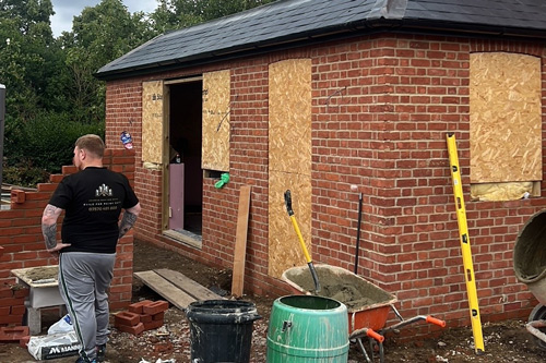 A construction worker is applying bricks to the exterior of a building under renovation, with plywood boards covering windows and doors, and construction tools and materials scattered around the site.