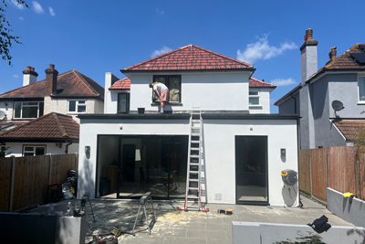 Construction worker on a ladder performing exterior work on a modern house with a flat roof; freshly painted white walls and a red tiled roof under a sunny sky.