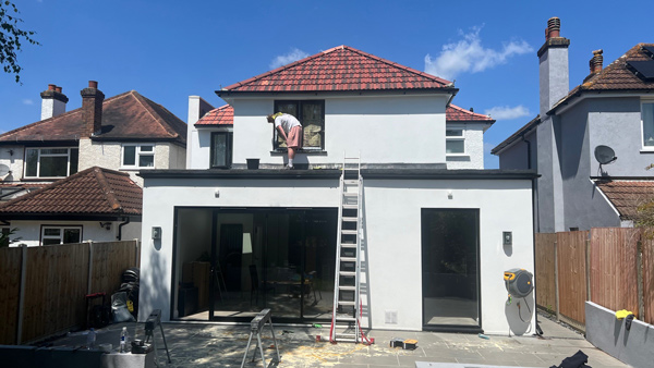 Worker renovating a house exterior while standing on a ladder, with a modern design featuring large windows and a tiled roof. Home improvement project in progress.